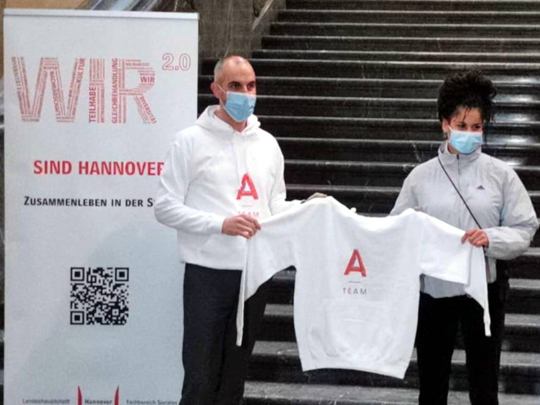Oberbürgermeister Belit Onay und eine Frau halten einen weißen Kapuzenpullover mit der Aufschrift A-Team. Sie stehen vor der Freitreppe im Neuen Rathaus. Links neben Onay steht ein Banner mit der Aufschrift "WIR 2.0 sind Hannover - Zusammnleben in der Stadt".